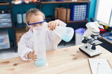 Adorable blonde girl student pouring liquid on test tube at laboratory classroom