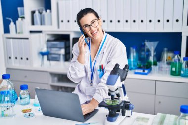 Young hispanic woman scientist talking on smartphone using laptop at laboratory