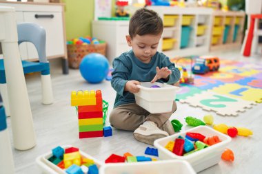 Adorable hispanic boy playing with construction blocks sitting on floor at kindergarten