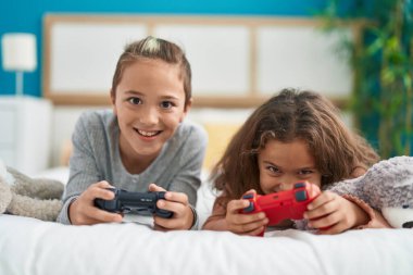 Two kids playing video game lying on bed at bedroom