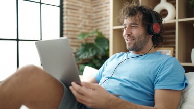 Young hispanic man using laptop and headphones lying on bed at bedroom