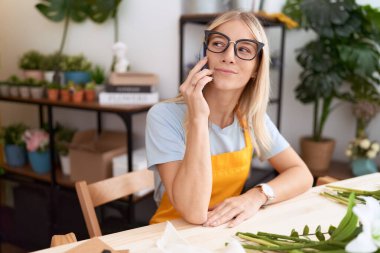 Young blonde woman florist smiling confident talking on smartphone at flower shop