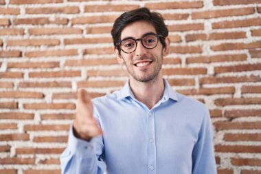 Young hispanic man standing over brick wall background smiling friendly offering handshake as greeting and welcoming. successful business. 