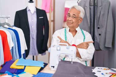 Middle age man with grey hair dressmaker using sewing machine pointing to both sides with fingers, different direction disagree 