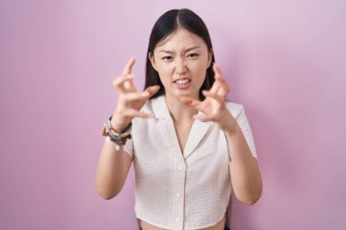 Chinese young woman standing over pink background shouting frustrated with rage, hands trying to strangle, yelling mad 