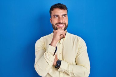 Handsome hispanic man standing over blue background with hand on chin thinking about question, pensive expression. smiling and thoughtful face. doubt concept. 