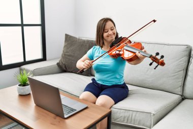 Down sendromlu esmer kadın kanepede oturup oturma odasında keman çalışıyor.