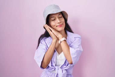 Young hispanic woman standing over pink background wearing hat sleeping tired dreaming and posing with hands together while smiling with closed eyes. 