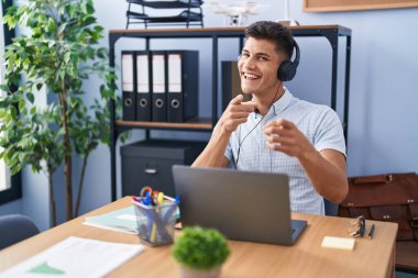 Young hispanic man working at the office wearing headphones pointing fingers to camera with happy and funny face. good energy and vibes. 