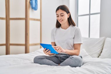 Young hispanic woman using touchpad sitting on bed at bedroom
