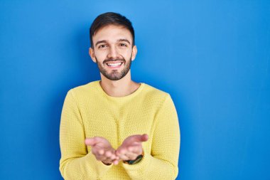 Hispanic man standing over blue background smiling with hands palms together receiving or giving gesture. hold and protection 