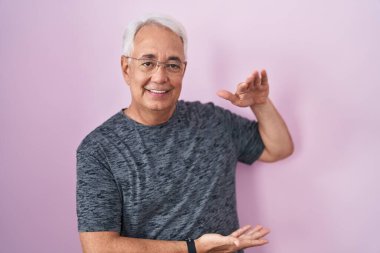 Middle age man with grey hair standing over pink background gesturing with hands showing big and large size sign, measure symbol. smiling looking at the camera. measuring concept. 