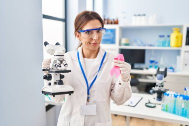 Young brunette woman working at scientist laboratory holding pink ribbon skeptic and nervous, frowning upset because of problem. negative person. 