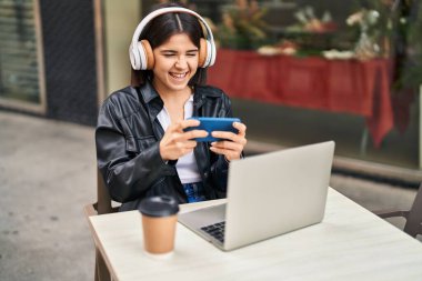 Young beautiful hispanic woman playing video game sitting on table at coffee shop terrace