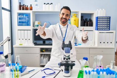 Young hispanic man with beard working at scientist laboratory looking at the camera smiling with open arms for hug. cheerful expression embracing happiness. 