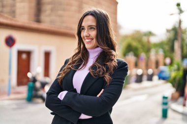 Genç İspanyol kadın gülümsüyor. Kollarını kavuşturup caddede duruyor.