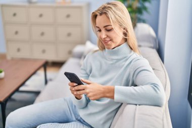 Evdeki kanepede oturan akıllı telefon kullanan genç sarışın kadın.