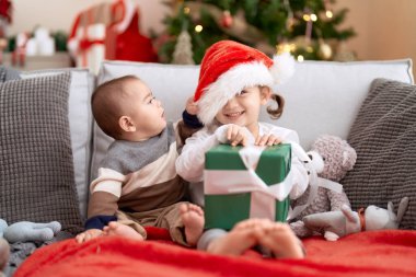 Brother and sister opening gift sitting on sofa by christmas tree at home