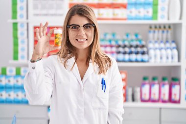 Young woman pharmacist smiling confident holding pills bottle at pharmacy