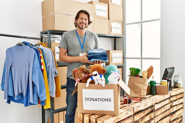 Кавказский мужчина средних лет улыбается уверенно держа сложенную одежду в благотворительном центре