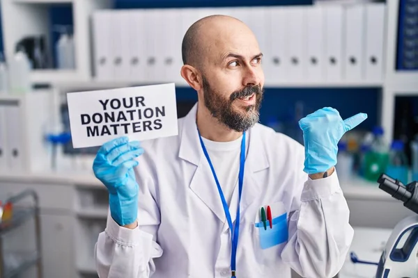 Young hispanic man working at scientist laboratory holding you donation matters banner pointing thumb up to the side smiling happy with open mouth