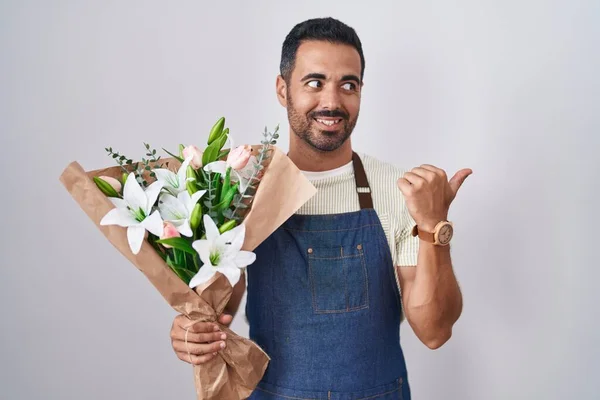 Hispanic man with beard working as florist smiling with happy face looking and pointing to the side with thumb up.