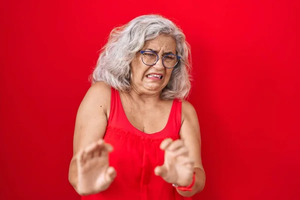 Middle age woman with grey hair standing over red background disgusted expression, displeased and fearful doing disgust face because aversion reaction.