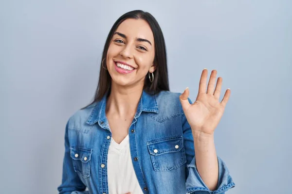 ヒスパニック系の女性が青の背景に立って手を振っこんにちは幸せと笑顔 優しい歓迎ジェスチャー — ストック写真