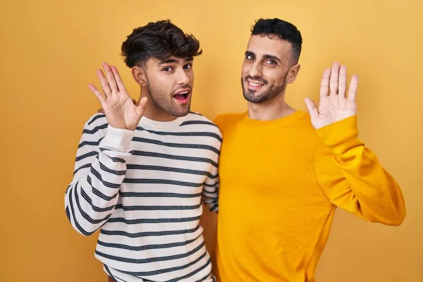 Young Hispanic Gay Couple Standing Yellow Background Waiving Saying Hello — Stok fotoğraf