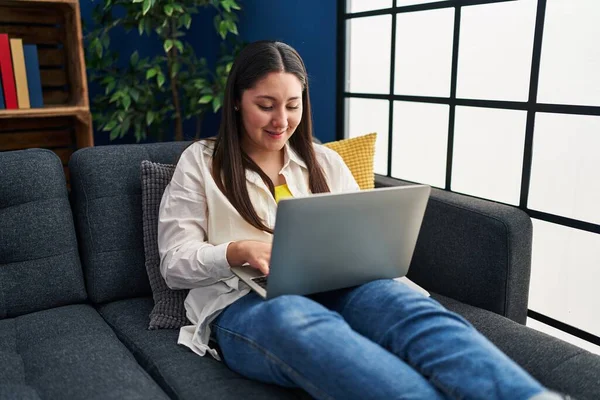 Genç İspanyol kadın evdeki koltukta dizüstü bilgisayar kullanıyor.