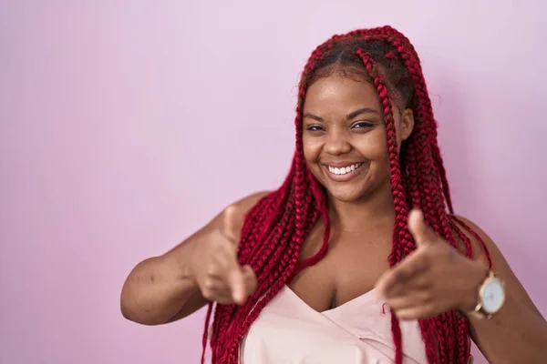 African american woman with braided hair standing over pink background pointing fingers to camera with happy and funny face. good energy and vibes.