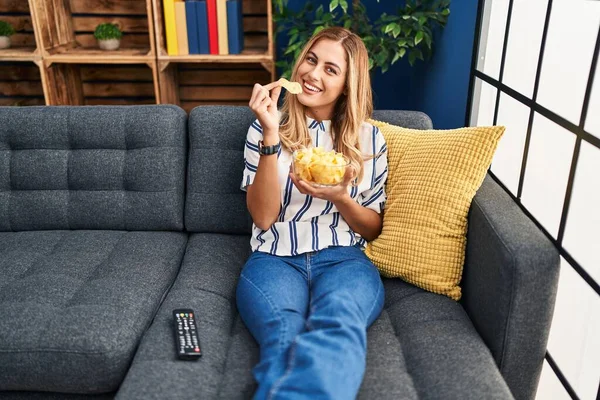 Young Blonde Woman Eating Chips Potatoes Sitting Sofa Home — Φωτογραφία Αρχείου