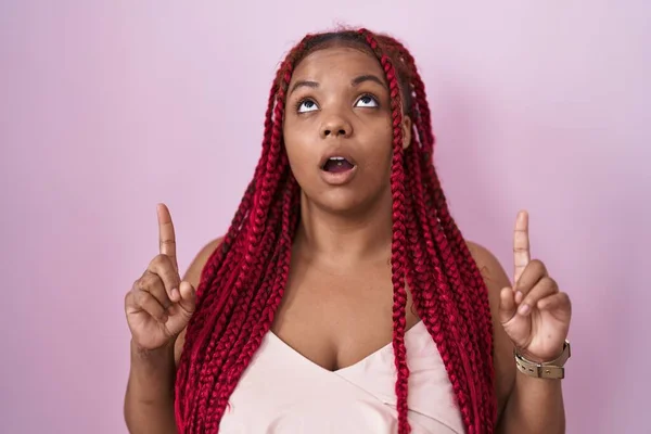African american woman with braided hair standing over pink background amazed and surprised looking up and pointing with fingers and raised arms.