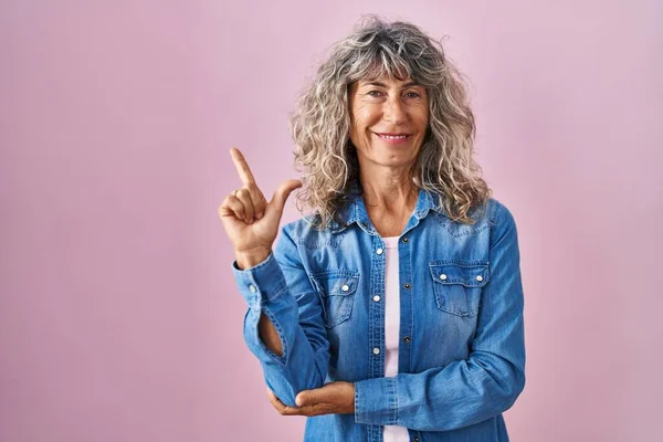 Middle age woman standing over pink background with a big smile on face, pointing with hand and finger to the side looking at the camera.