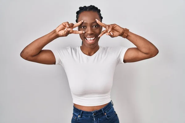 Beautiful black woman standing over isolated background doing peace symbol with fingers over face, smiling cheerful showing victory