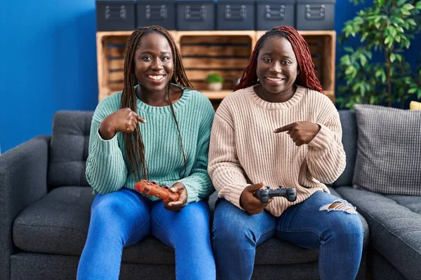 两个非洲女人在玩电子游戏 手里拿着控制器 指指点点着一个笑得开心而自豪的人 — 图库照片