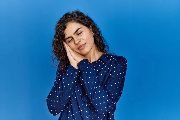 Young brunette woman with curly hair wearing casual clothes over blue background sleeping tired dreaming and posing with hands together while smiling with closed eyes.