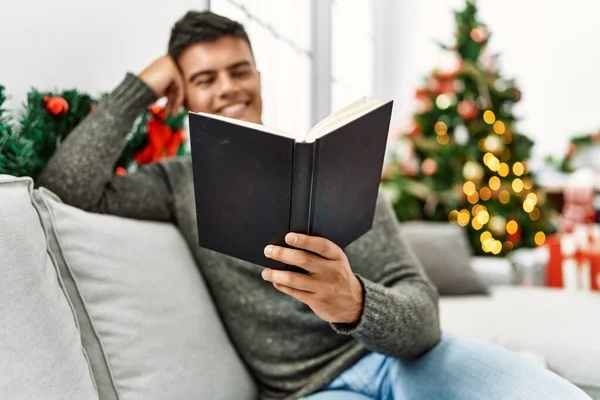 Young Hispanic Man Reading Book Sitting Sofa Christmas Tree Home — Stock Photo, Image