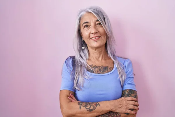 Middle age woman with tattoos standing over pink background happy face smiling with crossed arms looking at the camera. positive person.