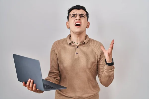 Non binary person using computer laptop crazy and mad shouting and yelling with aggressive expression and arms raised. frustration concept.