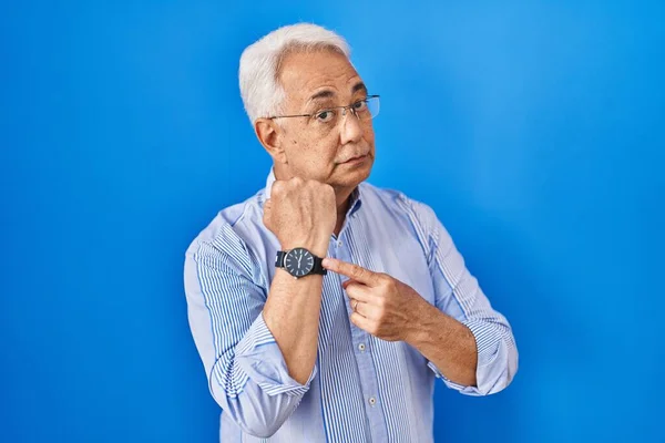Hispanic senior man wearing glasses in hurry pointing to watch time, impatience, looking at the camera with relaxed expression