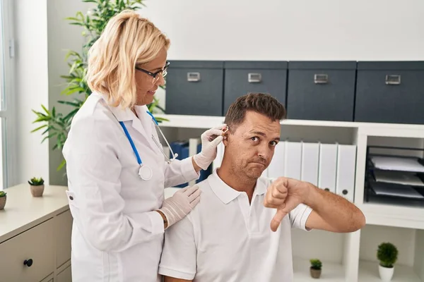 Hispanic man getting medical hearing aid at the doctor with angry face, negative sign showing dislike with thumbs down, rejection concept
