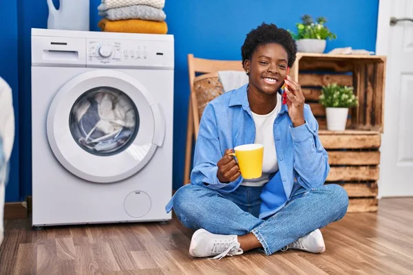 非洲裔美国妇女在洗衣房用智能手机聊天 喝咖啡等洗衣机 — 图库照片