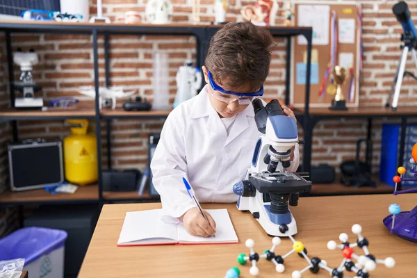 Adorable Hispanic Boy Student Using Microscope Writing Notebook Laboratory Classroom — Stockfoto