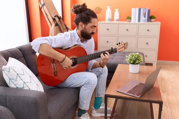 Young hispanic man having online guitar class sitting on sofa at home