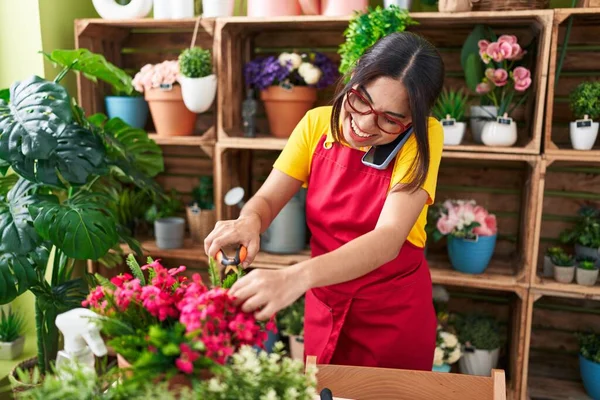 Young beautiful arab woman florist talking on smartphone cutting plant at flower shop