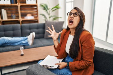Young hispanic woman working as psychology counselor crazy and mad shouting and yelling with aggressive expression and arms raised. frustration concept. 