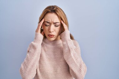Mavi arka planda duran İspanyol kadın, elleri başının üstünde, stres yüzünden baş ağrısı çekiyor. migren ağrısı. 