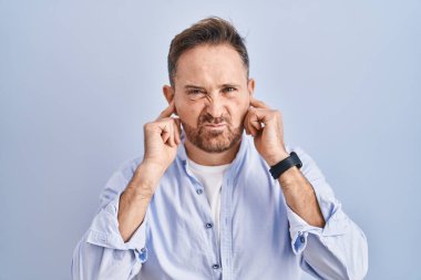 Middle age caucasian man standing over blue background covering ears with fingers with annoyed expression for the noise of loud music. deaf concept. 