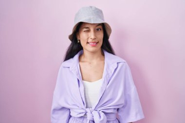 Young hispanic woman standing over pink background wearing hat winking looking at the camera with sexy expression, cheerful and happy face. 